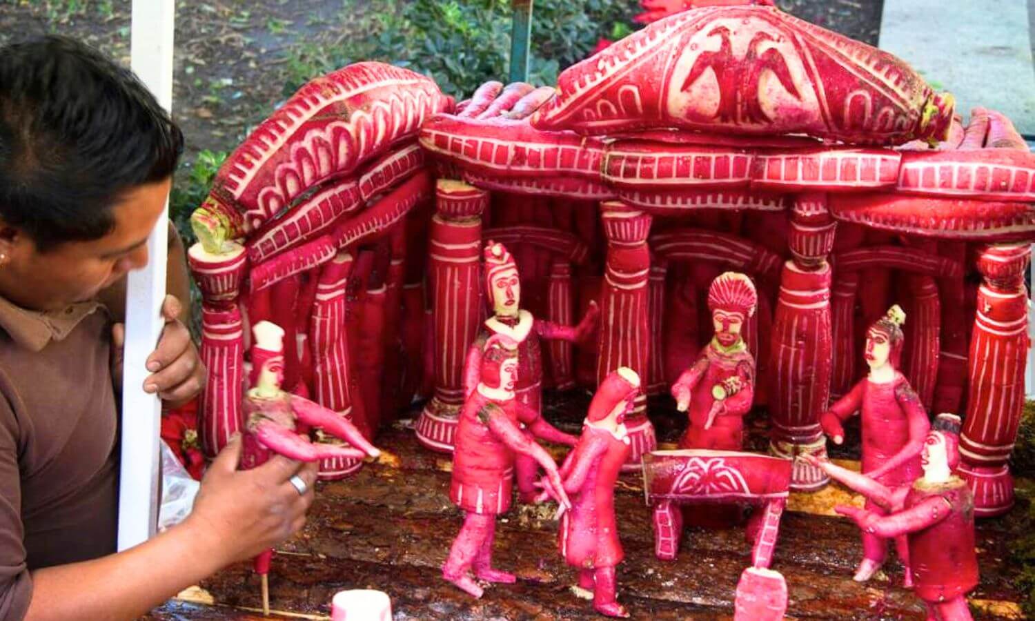 A man competing a scene with seven people in front of a pillared building entirely carved from radishes. The Noche de las Rábanos festival in Oaxaca city.