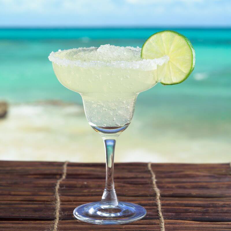 A frozen margarita in a salt rimmed glass on a table in front of the beach.