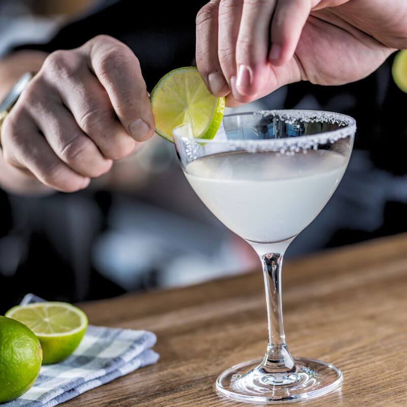 A man adding a slice of lime to the salt rimmed glass of margarita.