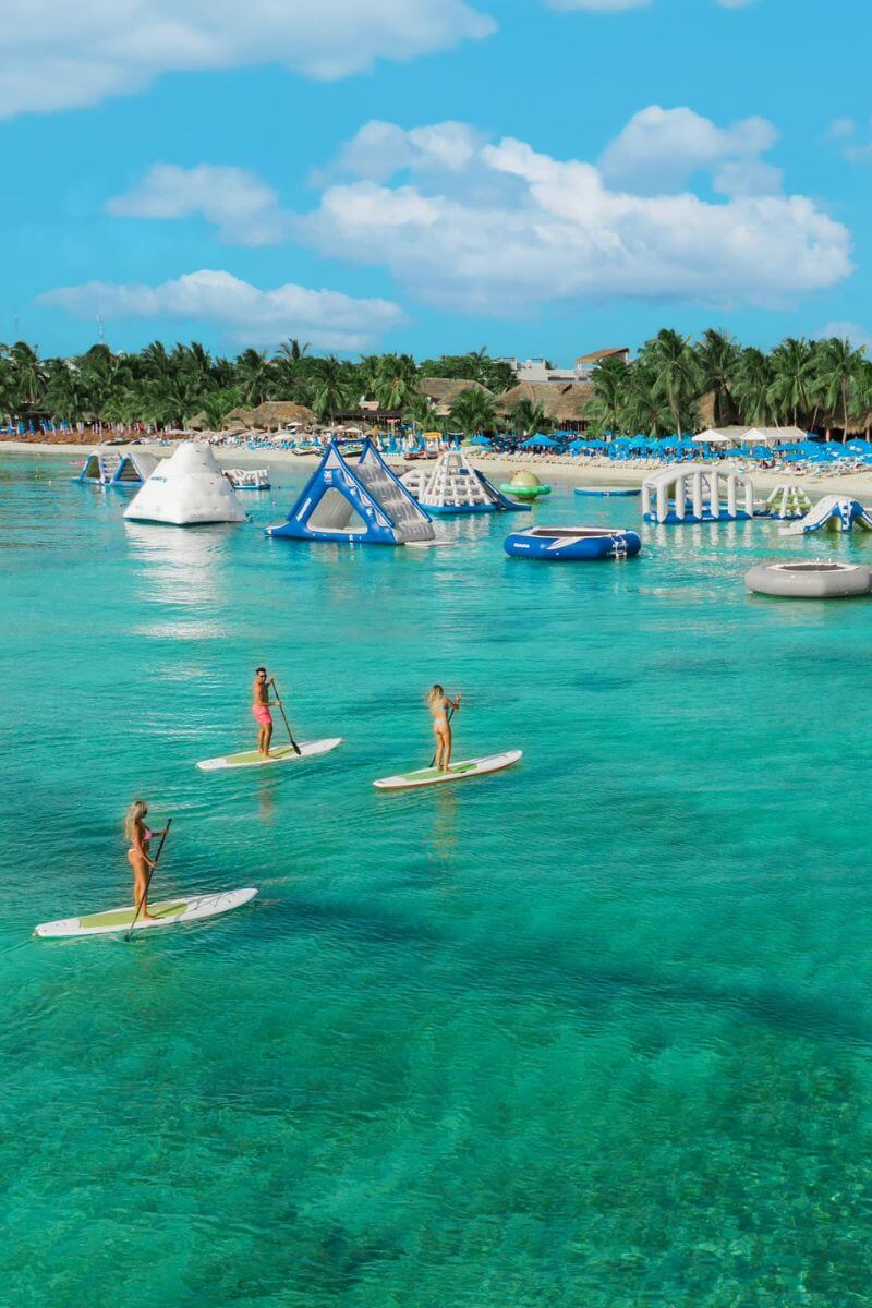 Three people on paddle boards in front fo the inflatable obstacle course at Paradise Beach, Cozumel.